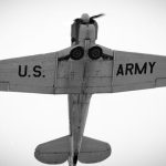 Black and white image capturing the underside of a vintage U.S. Army aircraft in flight.